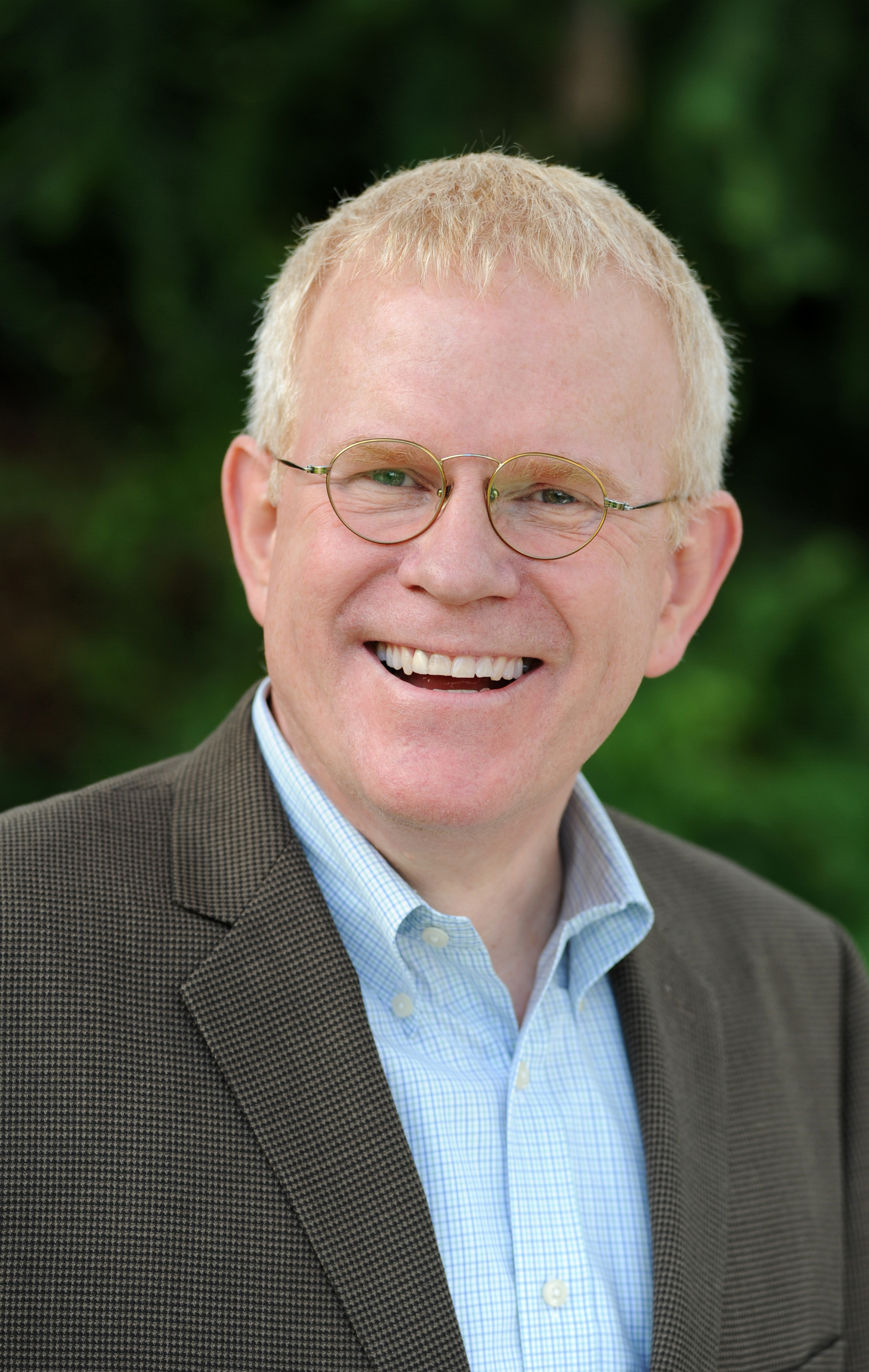 A man with light hair, glasses, and a big smile is wearing a suit jacket and a light blue collar shirt against a blurred green background, perhaps preparing to speak about Jurassic Parliament.