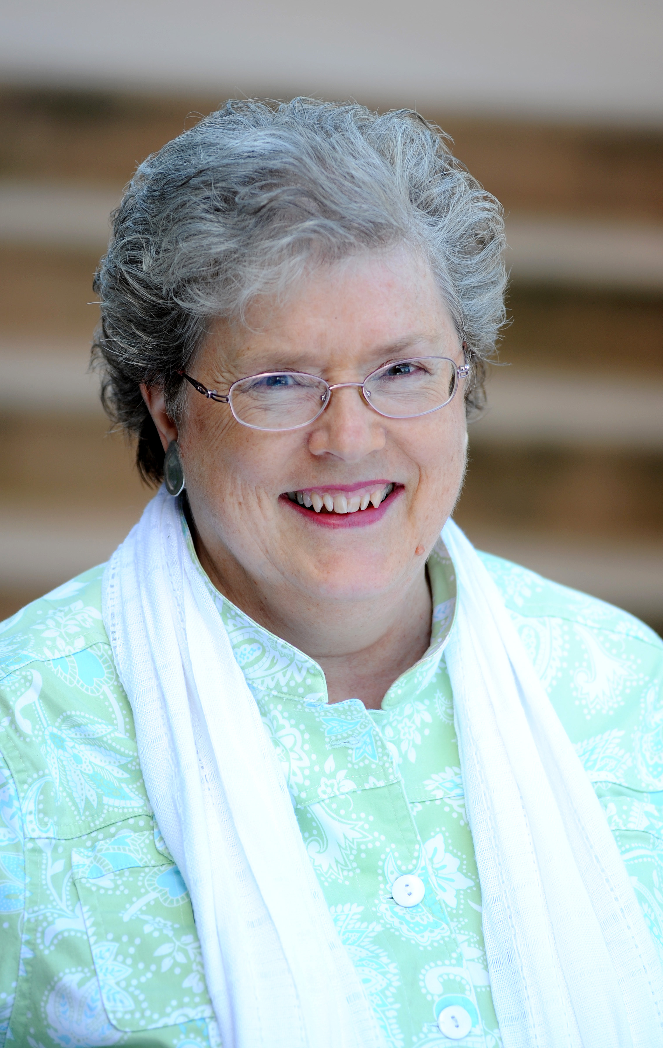 Elderly woman Ann Macfarlane, with short gray hair, glasses, and a green patterned shirt, smiling while standing in front of blurred stairs.