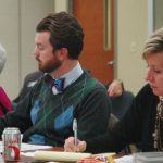 Three individuals sit at a conference table taking notes during a meeting. A can of soda and a snack bag are on the table, while one person looks visibly uncomfortable after inappropriate remarks were made.