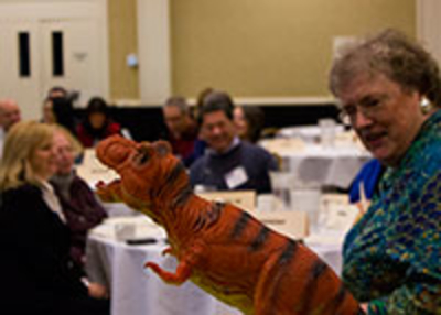 An older woman holds a toy dinosaur in front of a group of seated people in a conference room, sparking curiosity as she introduces the Roberts Rules online course.