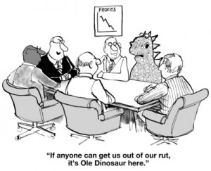 Cartoon of a business meeting with five men and a dinosaur around a table. A chart on the wall shows declining profits. One man raises his hand, asking questions about Robert's Rules. Caption reads, "If anyone can get us out of our rut, it’s Ole Dinosaur here.