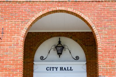 A brick archway frames a black lantern and a sign that reads "City Hall" above a white doorway, where the city's guidelines are upheld with the precision of Robert's Rules.