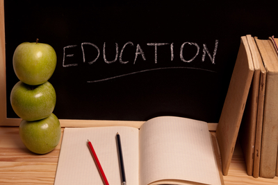 An open notebook with red and black pencils lies on a wooden table. Three green apples are stacked beside the notebook, reminiscent of Robert's Rules for orderly conduct. A blackboard with the word "EDUCATION" written in chalk is in the background.