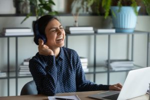 A woman wearing headphones smiles while sitting at a desk with a laptop and notepad in a home office setting, engaged in Robert's Rules training.