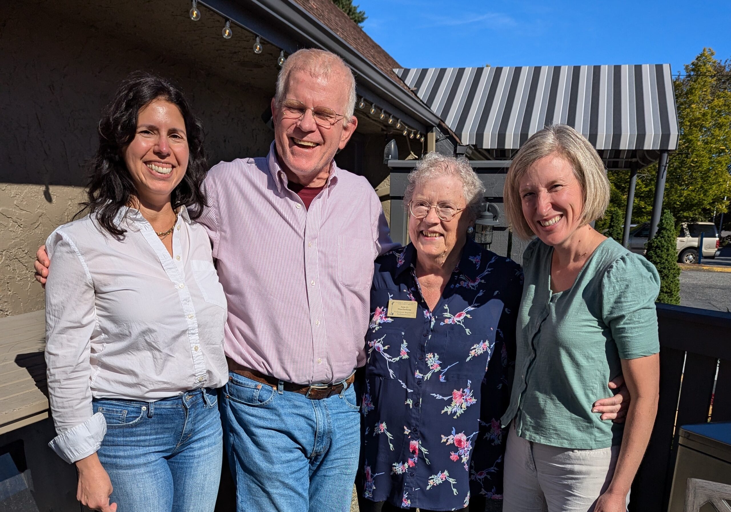 Four adults stand together outdoors, smiling at the camera. They are dressed casually, and a building with a striped awning is visible in the background.