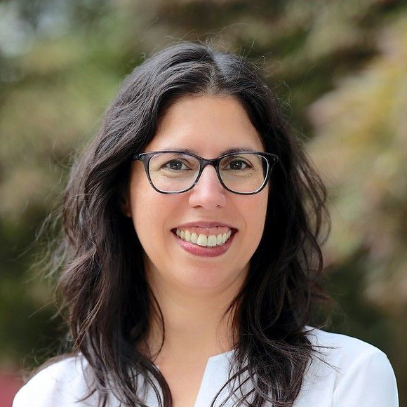 A woman with long dark hair, wearing glasses and a white shirt, smiles at the camera with a blurred outdoor background.