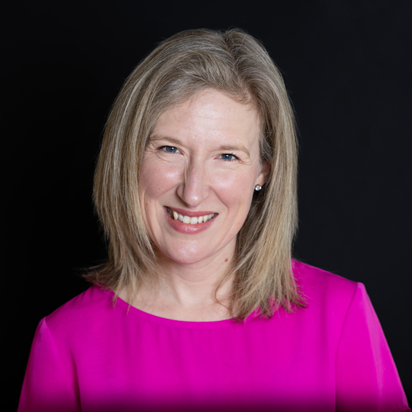 A woman with shoulder-length blonde hair smiles while wearing a bright pink top against a black background.