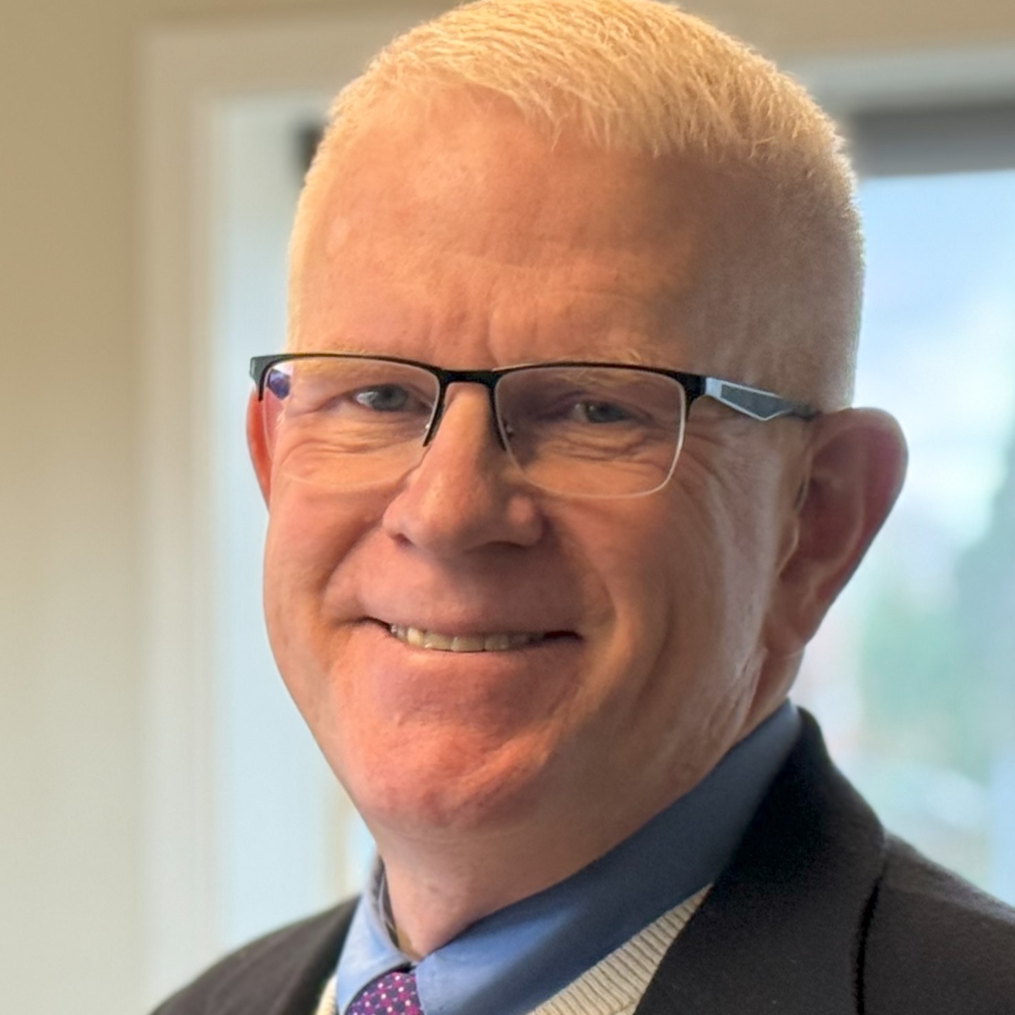 Andrew Estep, a man with short light hair and glasses, is smiling warmly indoors. He dons a blue shirt paired with a dark suit and patterned tie.