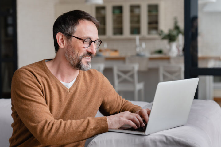 A man with glasses and a beard sits on a sofa using a laptop, smiling. He wears a brown sweater, and the background shows a kitchen with shelves and chairs.