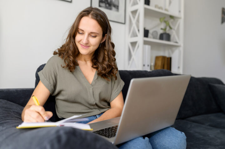 A woman sits on a couch with a laptop on her lap, smiling as she writes in a notebook. Behind her is a white bookshelf with decorations.