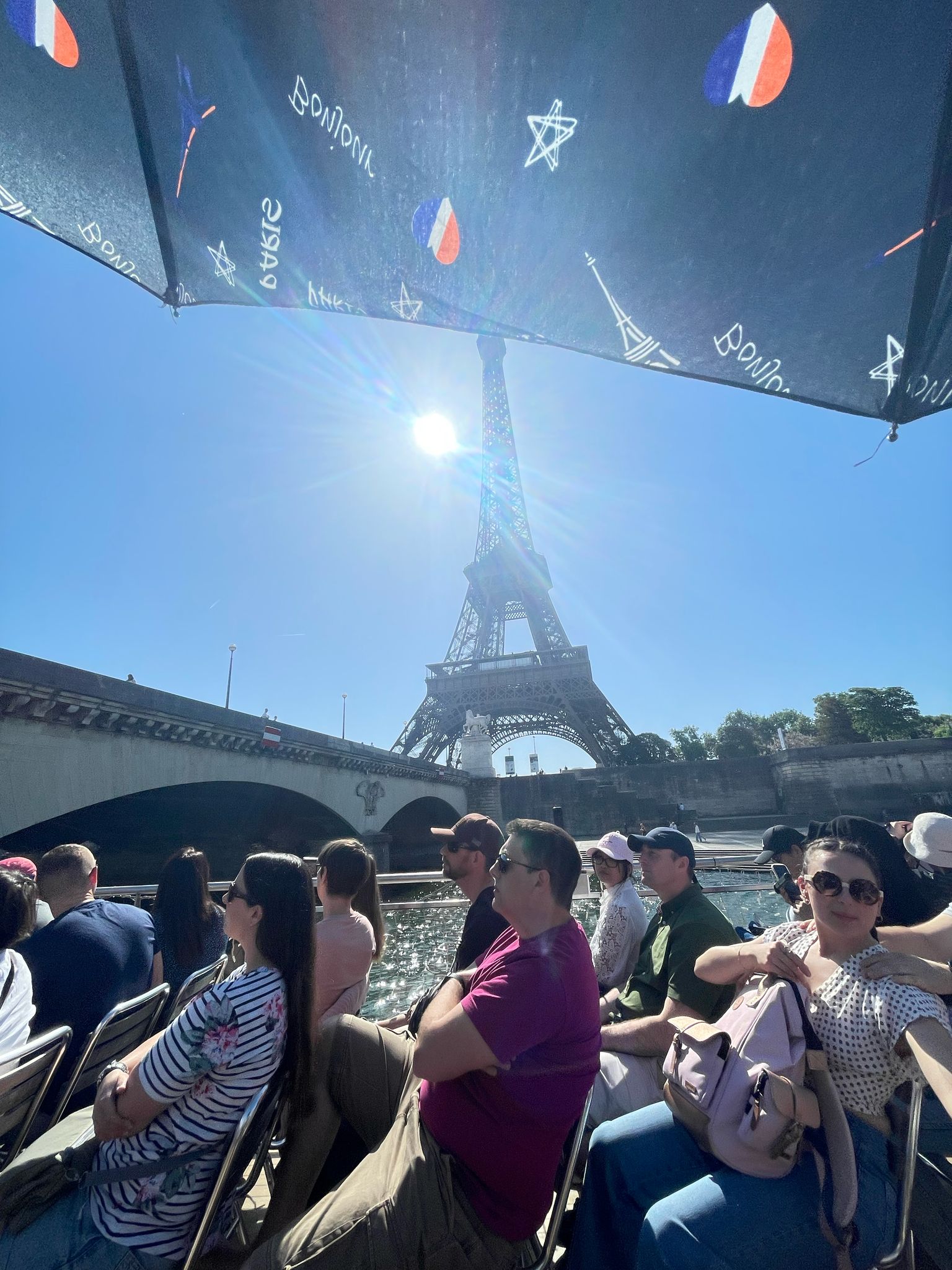 Tourists sit on a boat under a canopy with French flags and symbols, passing the Eiffel Tower on a sunny day in Paris.