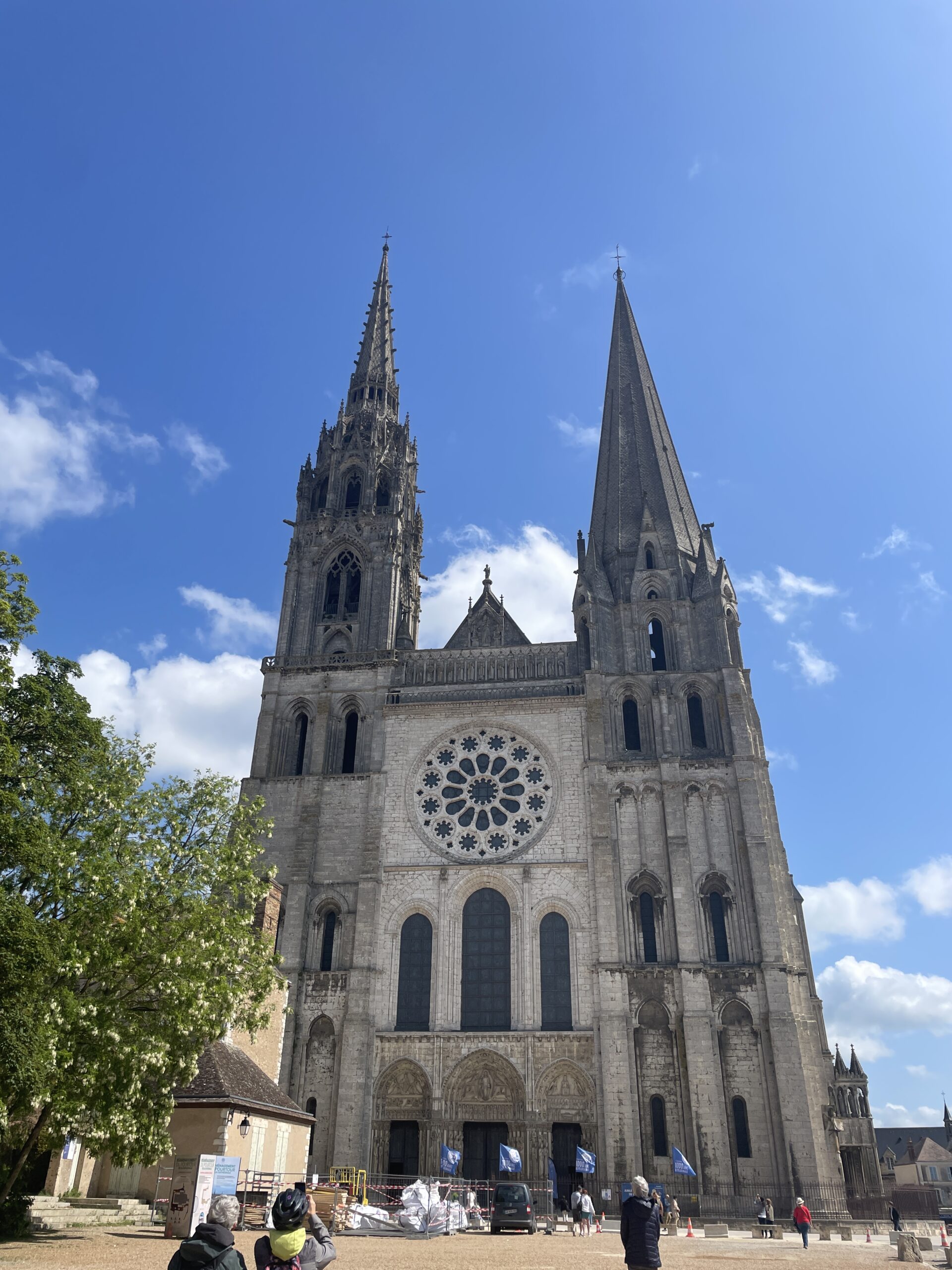 Front view of a large Gothic cathedral with two tall spires, a round rose window, and three arched entryways, under a blue sky with scattered clouds.