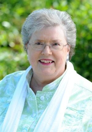 A smiling elderly woman with gray hair and glasses, wearing a light green patterned blouse and white scarf, stands in front of a blurred green background, ready to share her wisdom on Robert's Rules for elected officials.