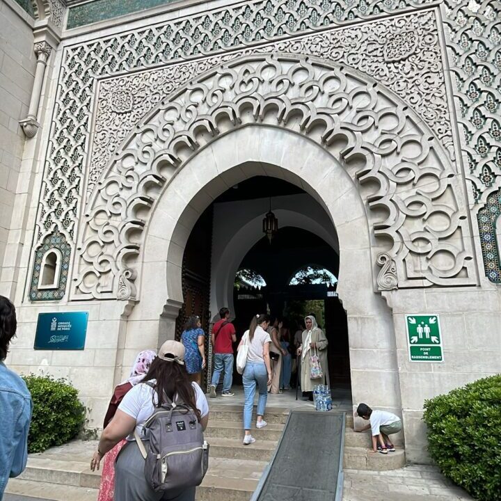 People enter a building with ornate geometric carvings above the arched entrance, a green star and crescent symbol on the roof, and a ramp leading to the doorway.