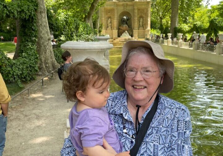 An older woman holding a toddler stands by a pond in a leafy park, with a stone fountain structure in the background.