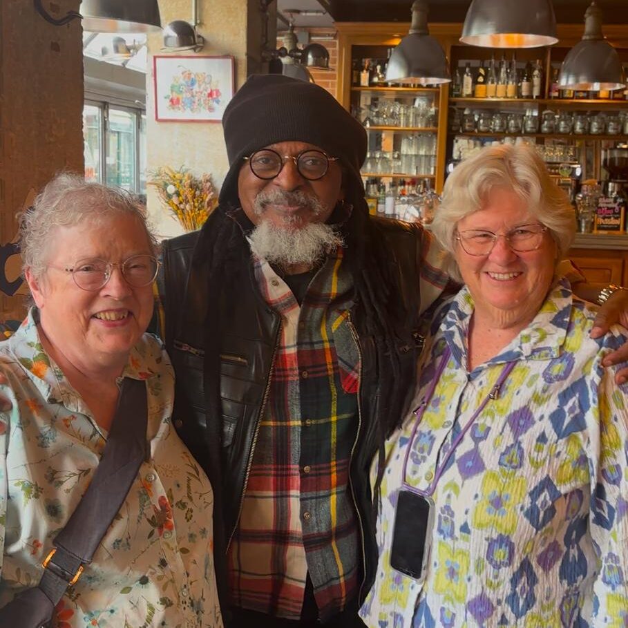 Three older adults, two women and one man, stand closely together and smile for a photo inside a bar or restaurant with shelves of bottles in the background.