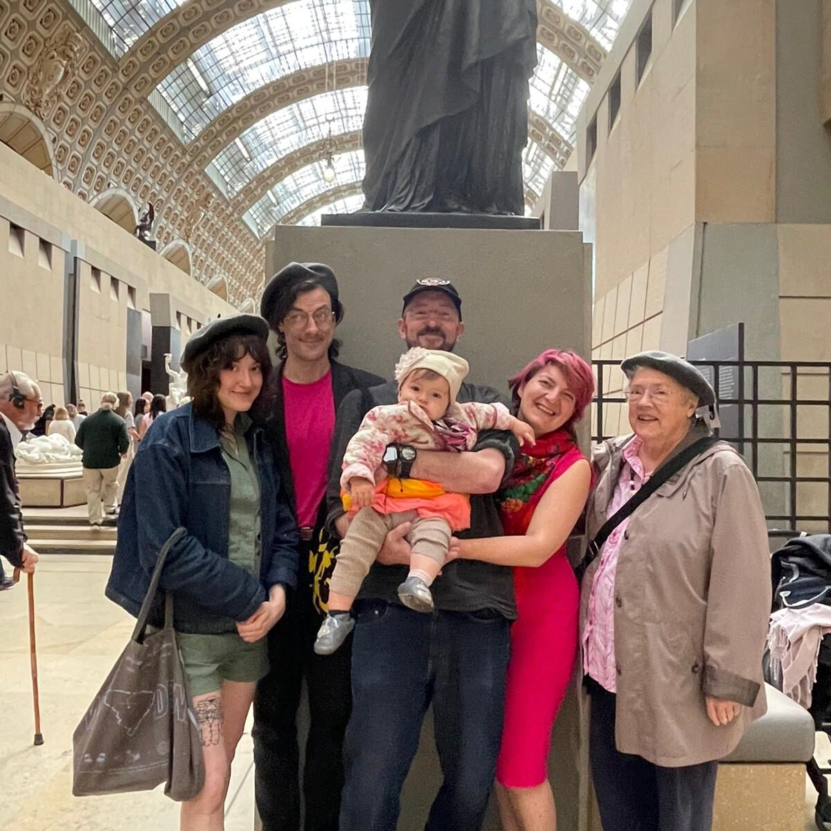 Six people, including a baby, pose together inside a large, ornate hall with a high glass ceiling and a statue in the background.