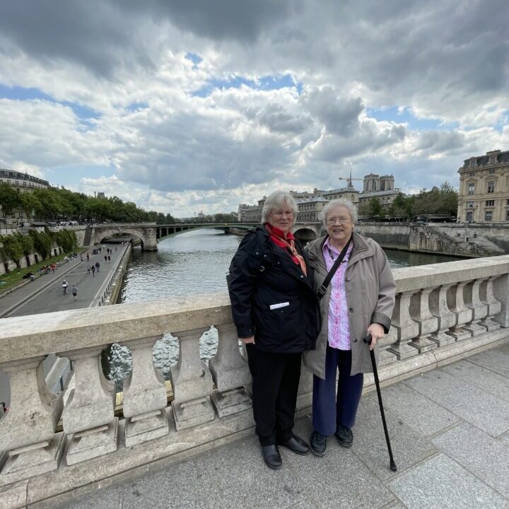 Two elderly women stand on a stone bridge over a river, with historic buildings and cloudy skies in the background.