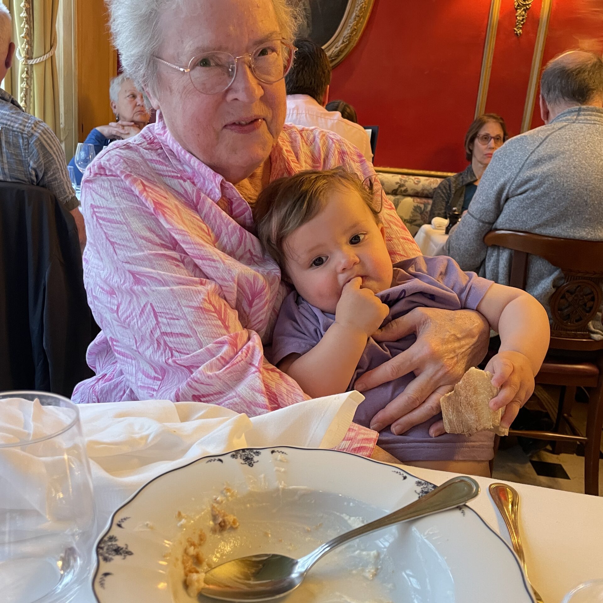 An elderly woman holds a young child on her lap at a restaurant table with an empty dessert plate in the foreground. Other diners are visible in the background.
