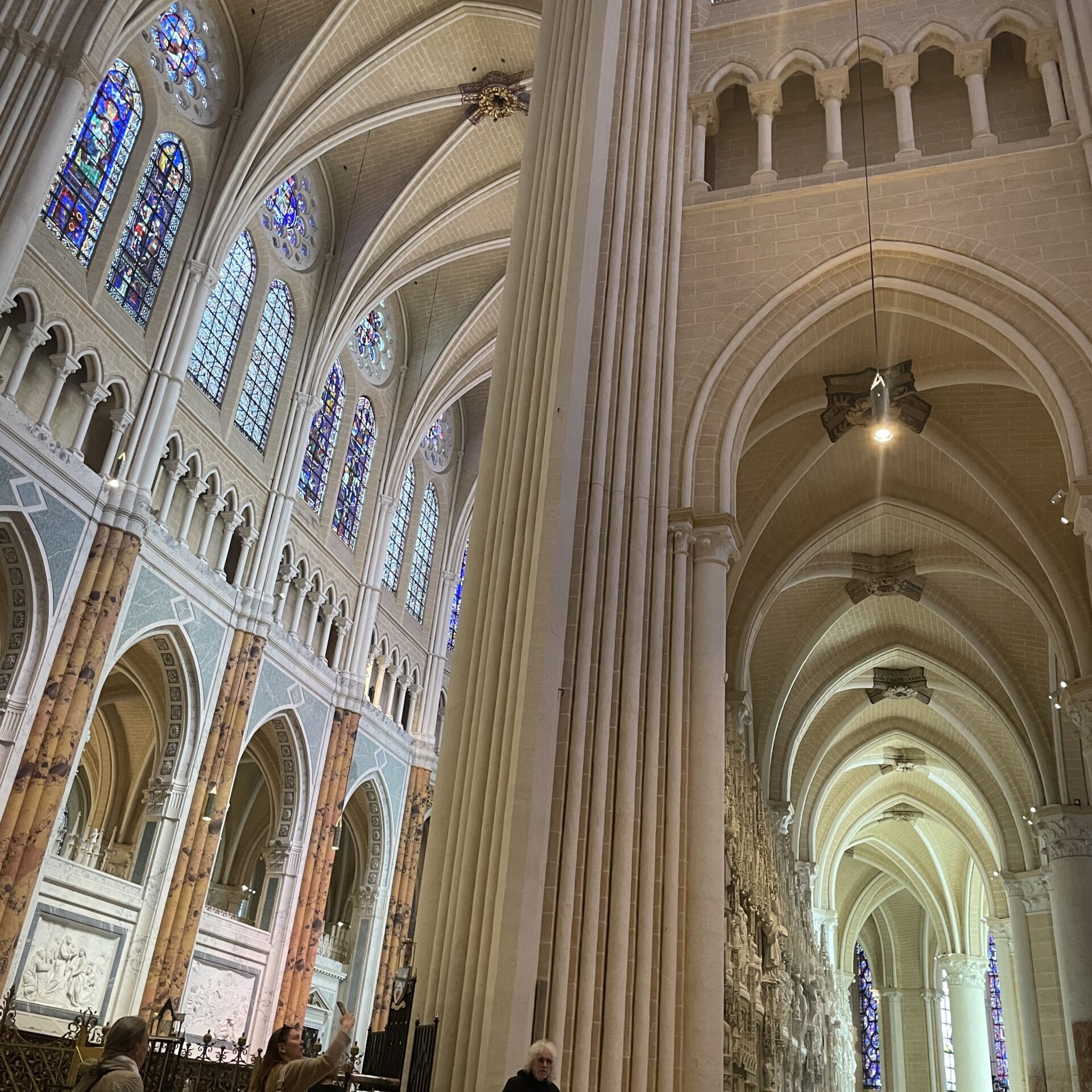 Interior of a grand cathedral with high vaulted ceilings, tall columns, stained glass windows, and a few people standing and sitting on wooden chairs.