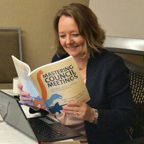 A woman sits at a desk with a laptop, smiling while reading a book titled "Mastering Council Meetings.