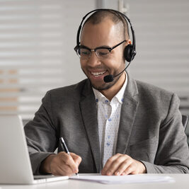 A man in a suit with glasses and a headset smiles while working on a laptop, writing notes as he engages in Robert's Rules training.