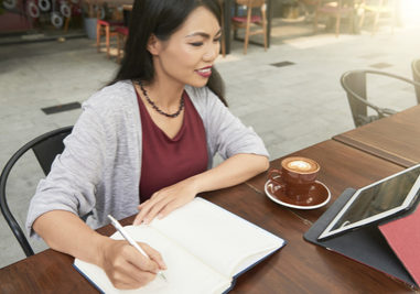 A woman is sitting at an outdoor table, writing in a notebook about Robert's Rules training, with a cup of coffee and a tablet beside her.