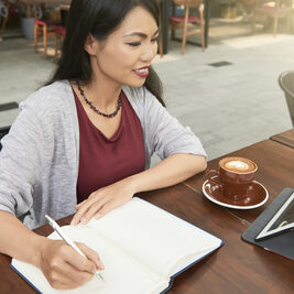 A woman is sitting at an outdoor table, writing in a notebook about Robert's Rules training, with a cup of coffee and a tablet beside her.